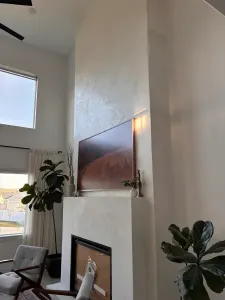 A modern living room with a tall white fireplace, a landscape photo above it, large windows, two armchairs, indoor plants, and sleek microtopping walls that add contemporary flair.