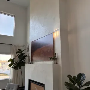 A modern living room with a tall white fireplace, a landscape photo above it, large windows, two armchairs, indoor plants, and sleek microtopping walls that add contemporary flair.