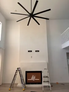A tall white fireplace with microtopping finishes and two wall mounts above it, a ladder and step stool in front, and a large black ceiling fan overhead in this modern living room.