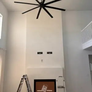 A tall white fireplace with microtopping finishes and two wall mounts above it, a ladder and step stool in front, and a large black ceiling fan overhead in this modern living room.
