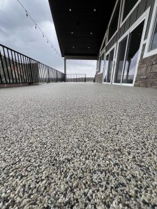 A low-angle view of a large, textured Resin Rock stone patio with glass doors, metal railings, and string lights under a covered roof.