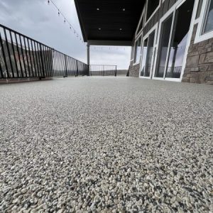 A low-angle view of a large, textured Resin Rock stone patio with glass doors, metal railings, and string lights under a covered roof.
