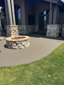 A circular stone fire pit sits on a concrete patio in front of a house with stone, wood, and resin rock exterior accents, right beside a grassy lawn.