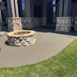 A circular stone fire pit sits on a concrete patio in front of a house with stone, wood, and resin rock exterior accents, right beside a grassy lawn.