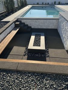 Modern outdoor patio with a rectangular fire pit surrounded by black stones and resin rock, a wooden bench, steps, and a swimming pool in the background.