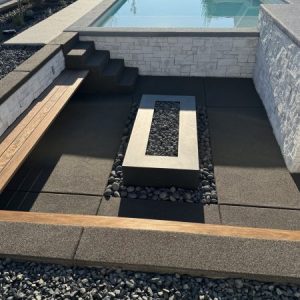 Modern outdoor patio with a rectangular fire pit surrounded by black stones and resin rock, a wooden bench, steps, and a swimming pool in the background.
