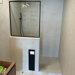 Minimalist walk-in shower with a black-framed glass divider, overhead rain shower, wall-mounted black toilet paper holder, and beige microtopping walls and floor.