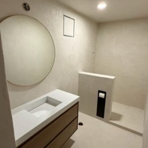 Minimalist bathroom with microtopping walls, a round mirror above a floating vanity, built-in sink, paper holder, and an open showers area—all in a neutral color scheme.