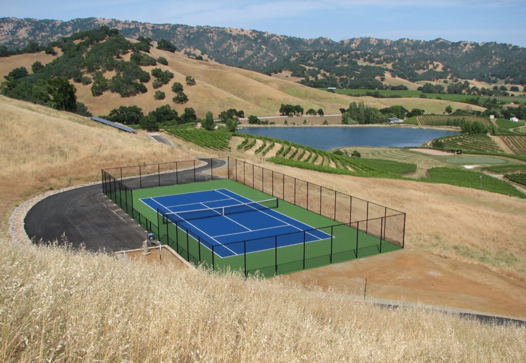 A tennis court with a blue and green surface is gracefully perched on a hillside, its vibrancy contrasting against the dry grass. Discreet concrete stains add character to the scene, while a shimmering lake and expansive vineyards complete the picturesque backdrop. A tennis court with a blue and green surface is gracefully perched on a hillside, its vibrancy contrasting against the dry grass. Discreet concrete stains add character to the scene, while a shimmering lake and expansive vineyards complete the picturesque backdrop.