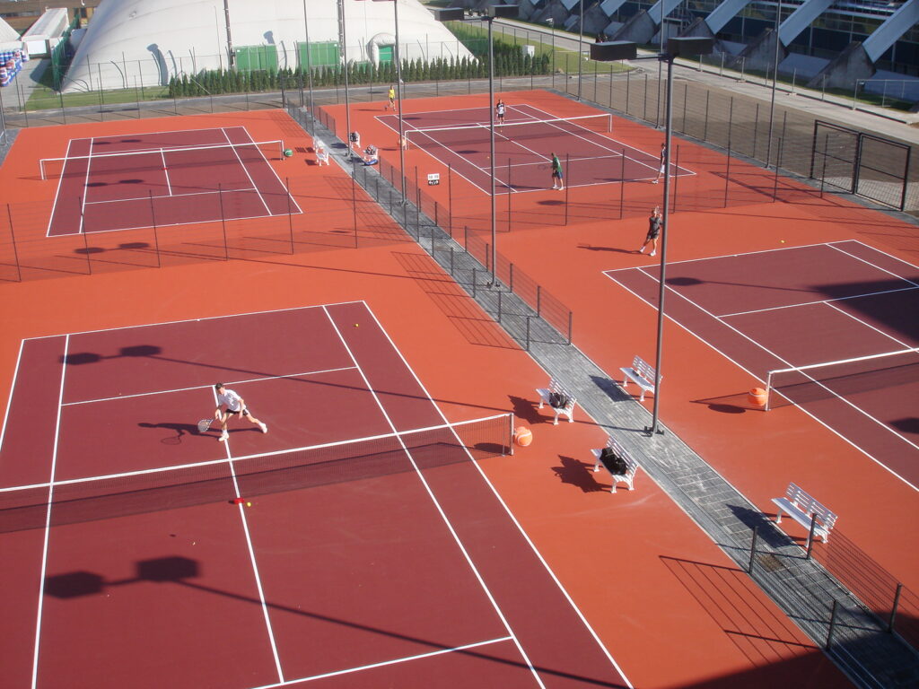 Aerial view of an outdoor tennis court complex with four courts, their surfaces marked by subtle concrete stains. Several people are playing or practicing tennis under clear skies. Aerial view of an outdoor tennis court complex with four courts, their surfaces marked by subtle concrete stains. Several people are playing or practicing tennis under clear skies.