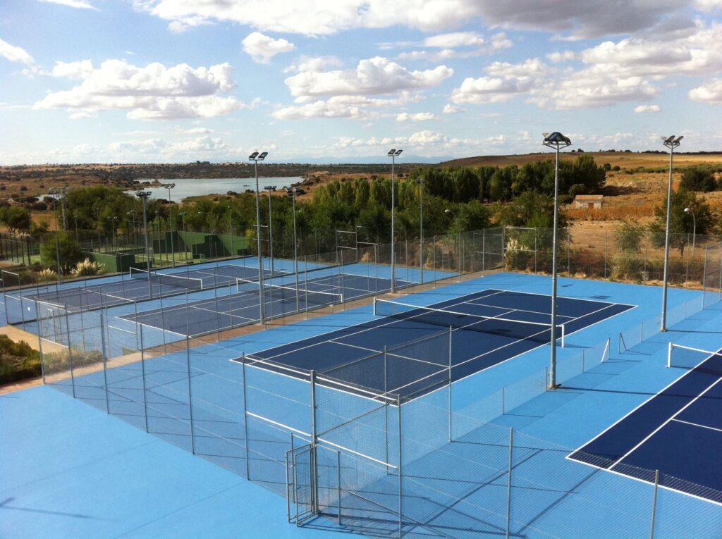 Outdoor tennis courts with blue surfaces and subtle concrete stains are surrounded by wire fences. They rest in a rural area where trees frame the scene, water shimmers in the background, and partly cloudy skies stretch above. Outdoor tennis courts with blue surfaces and subtle concrete stains are surrounded by wire fences. They rest in a rural area where trees frame the scene, water shimmers in the background, and partly cloudy skies stretch above.