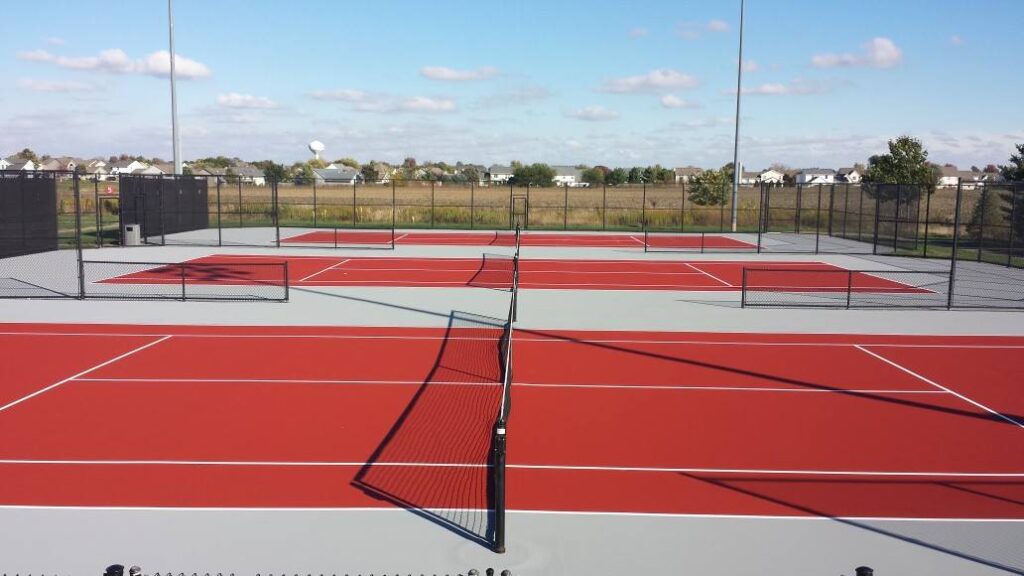 Four outdoor pickleball courts with red playing surfaces and gray borders, surrounded by black fencing, lie under a partly cloudy sky, enhanced by concrete stains that add character and depth to the surrounding walkway. Four outdoor pickleball courts with red playing surfaces and gray borders, surrounded by black fencing, lie under a partly cloudy sky, enhanced by concrete stains that add character and depth to the surrounding walkway.
