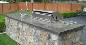 Outdoor kitchen featuring a built-in stainless steel grill and small sink on a stone countertop with stain concrete color accents. Surrounded by lush grass, a wooden fence, and an inviting patio.