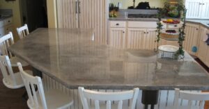 A kitchen island with a polished concrete countertop in a warm hue, surrounded by four white chairs, and a tiered stand holding fruit and greenery.