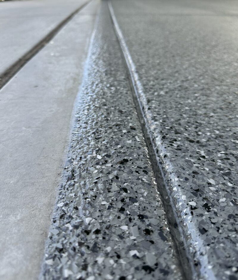 Close-up of a concrete floor coated with Coatary Epoxy Flakes, featuring a speckled finish and two narrow grooves parallel to a smooth, lighter section of concrete.