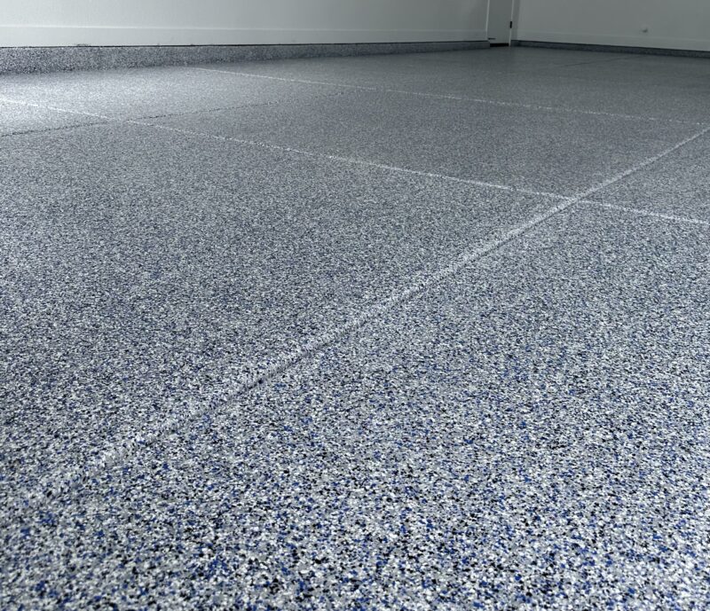 Close-up of a smooth, gray and blue Coatary Epoxy Flakes-coated floor in an empty room with white walls.