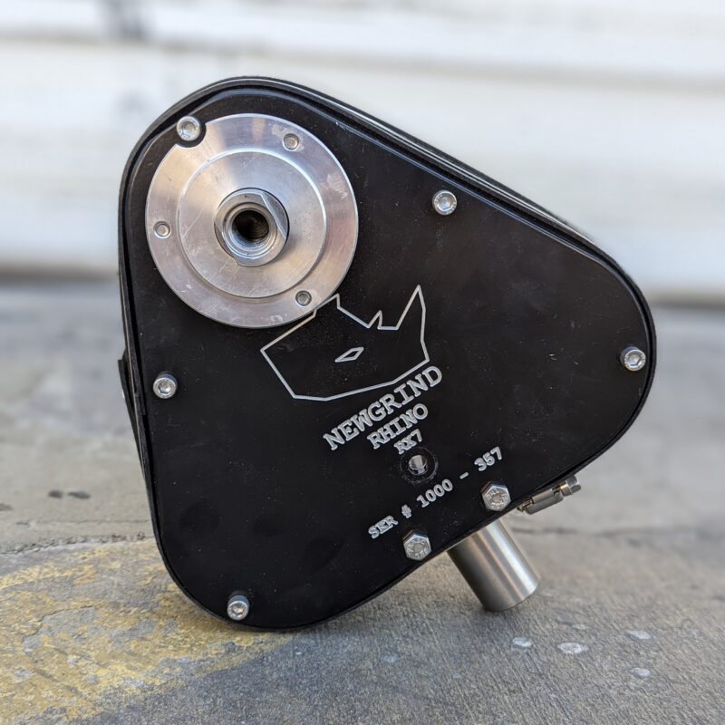 A black metal gearbox for the NewGrind RX7 Mini - 3 Head Grinder, with a top metal mounting point and a bottom cylindrical shaft, is shown on concrete.