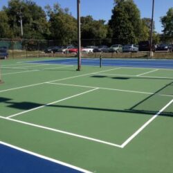 Empty outdoor tennis courts with green and blue surfaces, recently resurfaced by NovaSURFACE Resurfacer, are enclosed by a chain-link fence. Trees and parked cars appear in the background beneath a clear blue sky.
