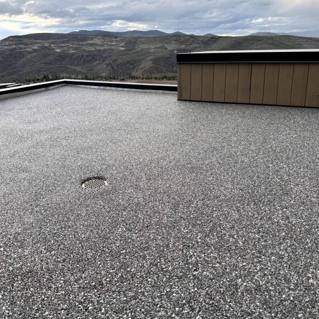 Flat gravel rooftop featuring Resin Rock with a drainage grate and a short brown wall in the background, overlooking distant hills under a cloudy sky.
