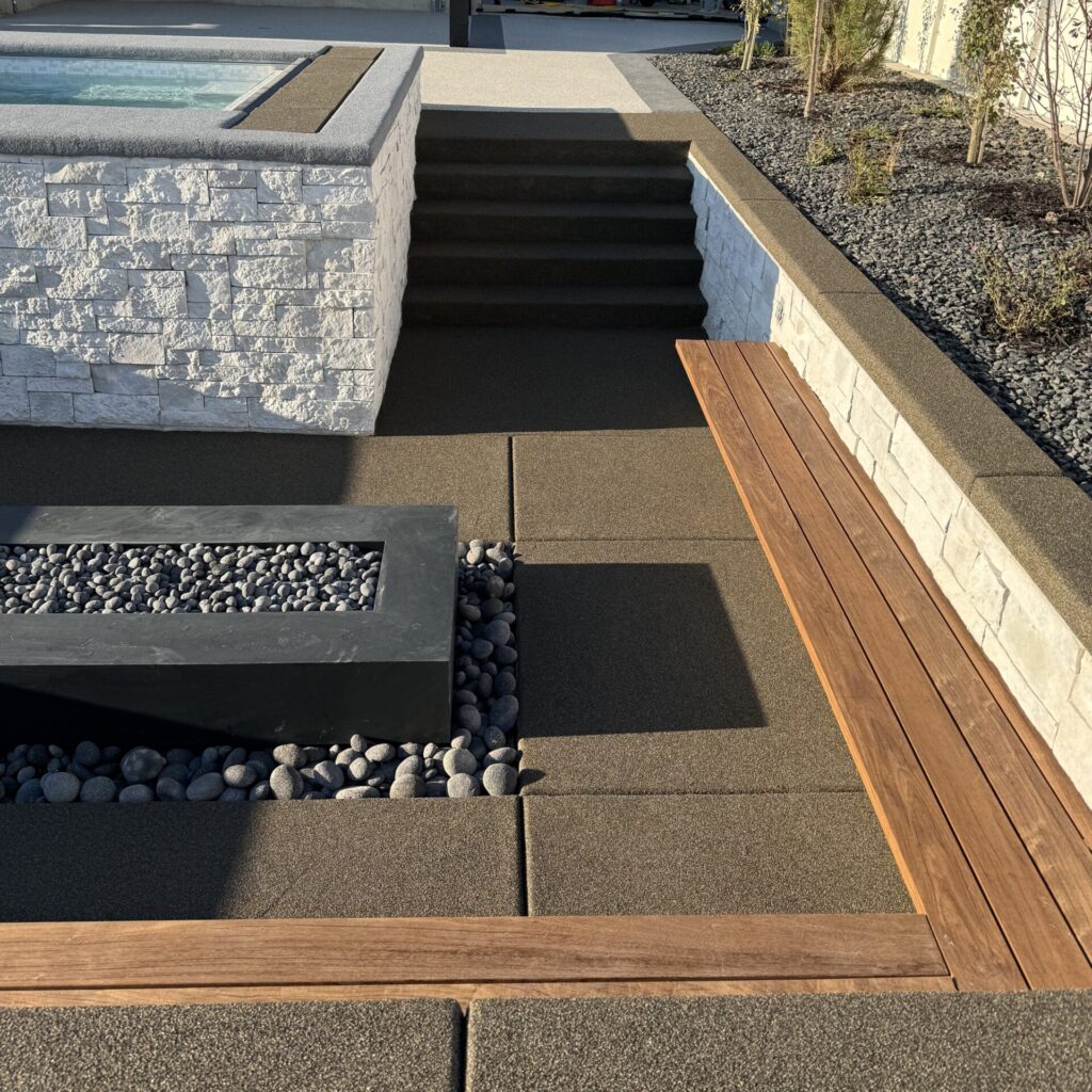 Modern backyard with a small pool, stone steps, a wooden bench, landscaped rocks, and workers in the background near a house. Resin Rock accents add a contemporary touch to the stylish outdoor space.