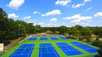 Aerial view of multiple blue and green tennis courts resurfaced with NovaSURFACE Resurfacer, surrounded by trees on a clear day with scattered clouds. Aerial view of multiple blue and green tennis courts resurfaced with NovaSURFACE Resurfacer, surrounded by trees on a clear day with scattered clouds.