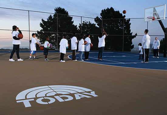 People lined up on an outdoor basketball court, each holding a basketball above the concrete stains, with a marked logo proudly displayed on the ground. Trees and a chain-link fence form the backdrop. People lined up on an outdoor basketball court, each holding a basketball above the concrete stains, with a marked logo proudly displayed on the ground. Trees and a chain-link fence form the backdrop.