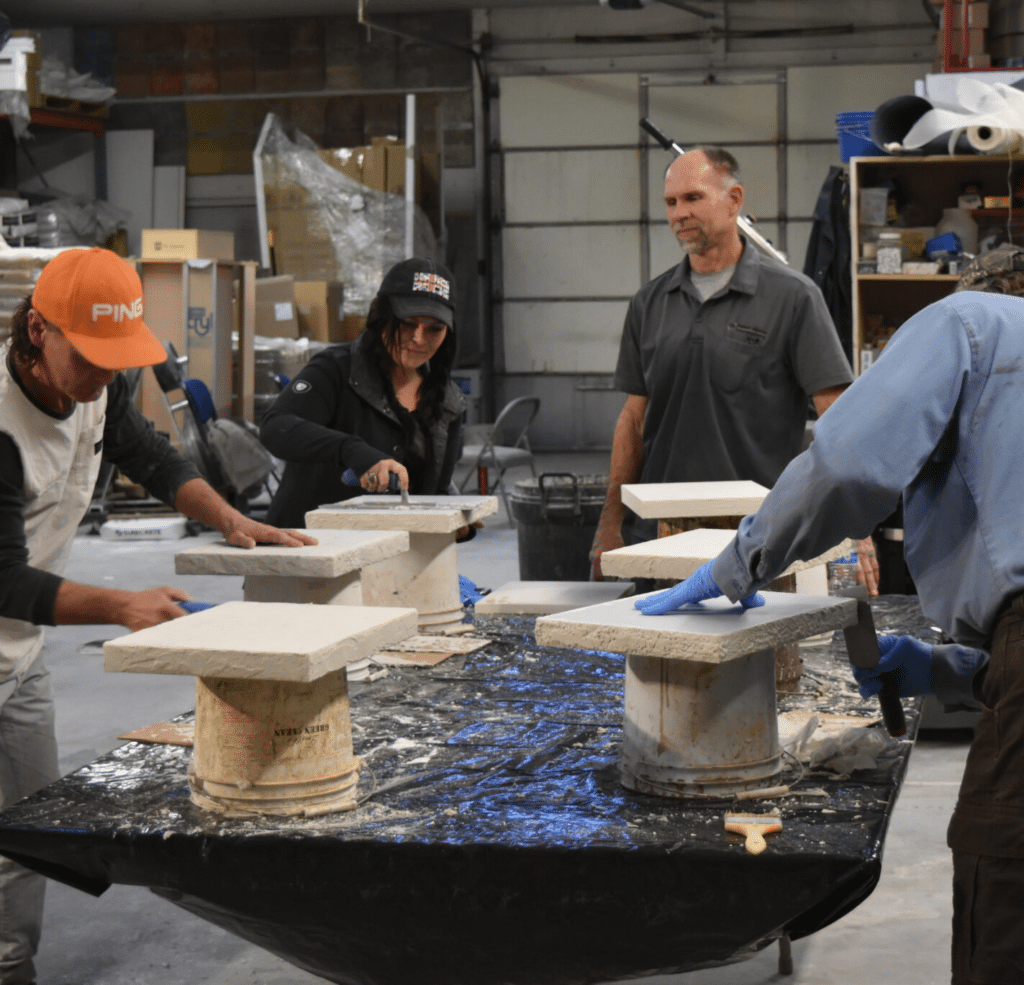 People working collaboratively on stone crafting projects in a workshop, with concrete overlays adding texture and depth to their creations. Tools and materials are neatly arranged on the table.