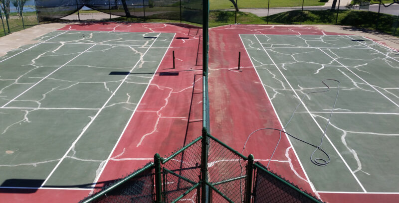 Two cracked and worn tennis courts with faded lines and missing nets, encircled by a green fence and lush greenery, await NovaSURFACE Resurfacer's transformative touch for a beautiful refresh.